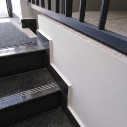 Zbigniew Grożyński Gro-Bud Usług remontowo budowlane - Fragment klatki schodowej z ciemnymi, granitowymi schodami z trzema jasnymi paskami, jasną ścianą z widocznymi drobnymi niedoskonałościami w postaci zacieków i ciemną metalową barierką.