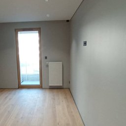 Zbigniew Grożyński Gro-Bud Usług remontowo budowlane - Wnętrze po odświeżeniu: gładkie, szare ściany, jasna drewniana podłoga, biały grzejnik pod oknem, widoczne gniazdka elektryczne.
