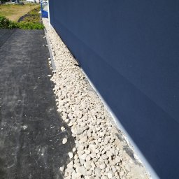 Zbigniew Grożyński Gro-Bud Usług remontowo budowlane - Świeżo pomalowana, gładka, ciemnoniebieska ściana budynku z ułożonym pasem białych kamieni dekoracyjnych wzdłuż jej podstawy, oddzielonych czarną geowłókniną od trawnika.
