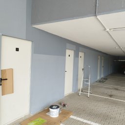 Zbigniew Grożyński Gro-Bud Usług remontowo budowlane - Korytarz garażowy z pomalowanymi ścianami na szaro-niebieski kolor, białe drzwi z numerami, drabina aluminiowa, puszka farby na kartonie i fragment zabezpieczonej kartonem klamki.