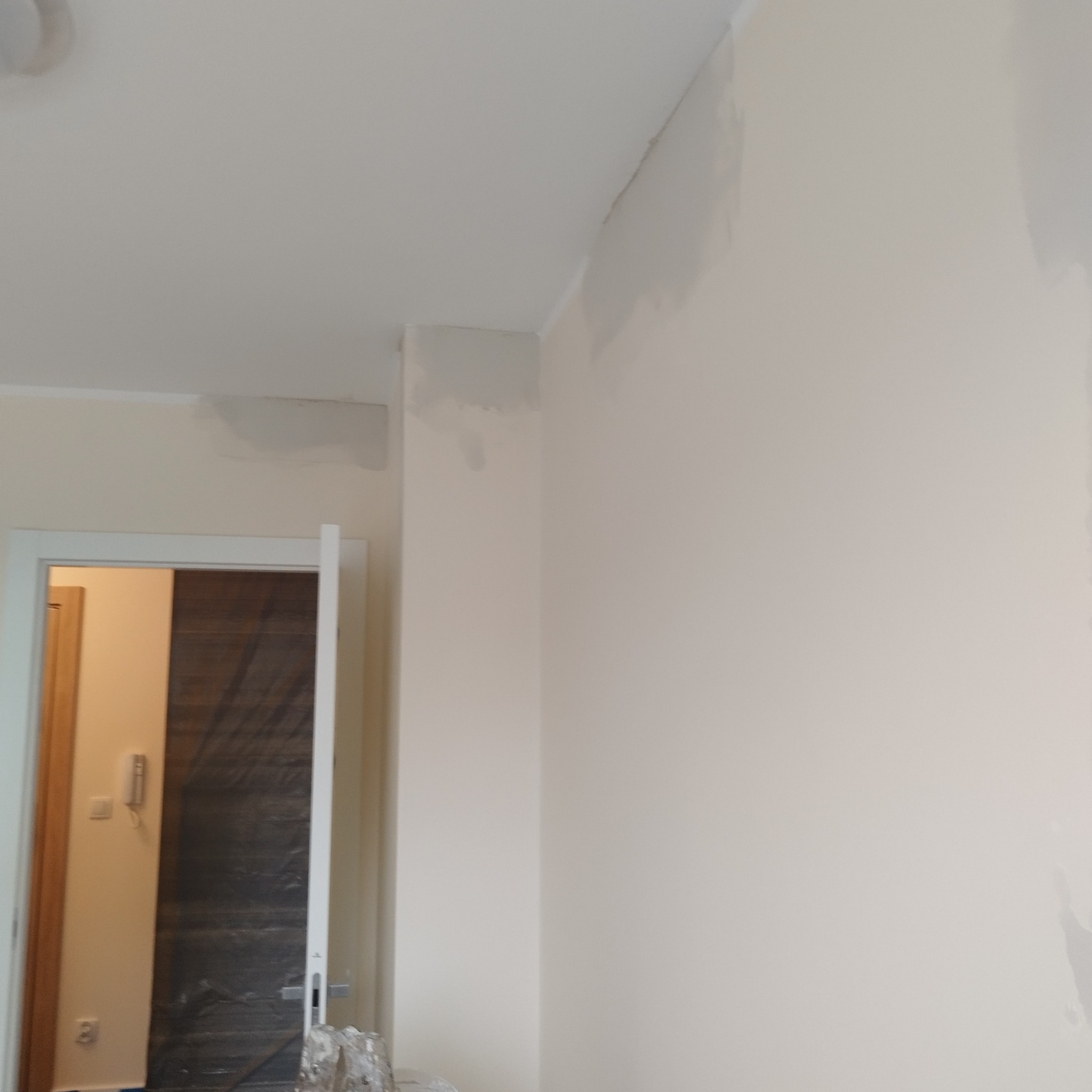 Ściana w trakcie prac wykończeniowych, widoczne ślady po szpachlowaniu i przygotowaniu do malowania, fragment otwartej drzwi z folią ochronną w tle.