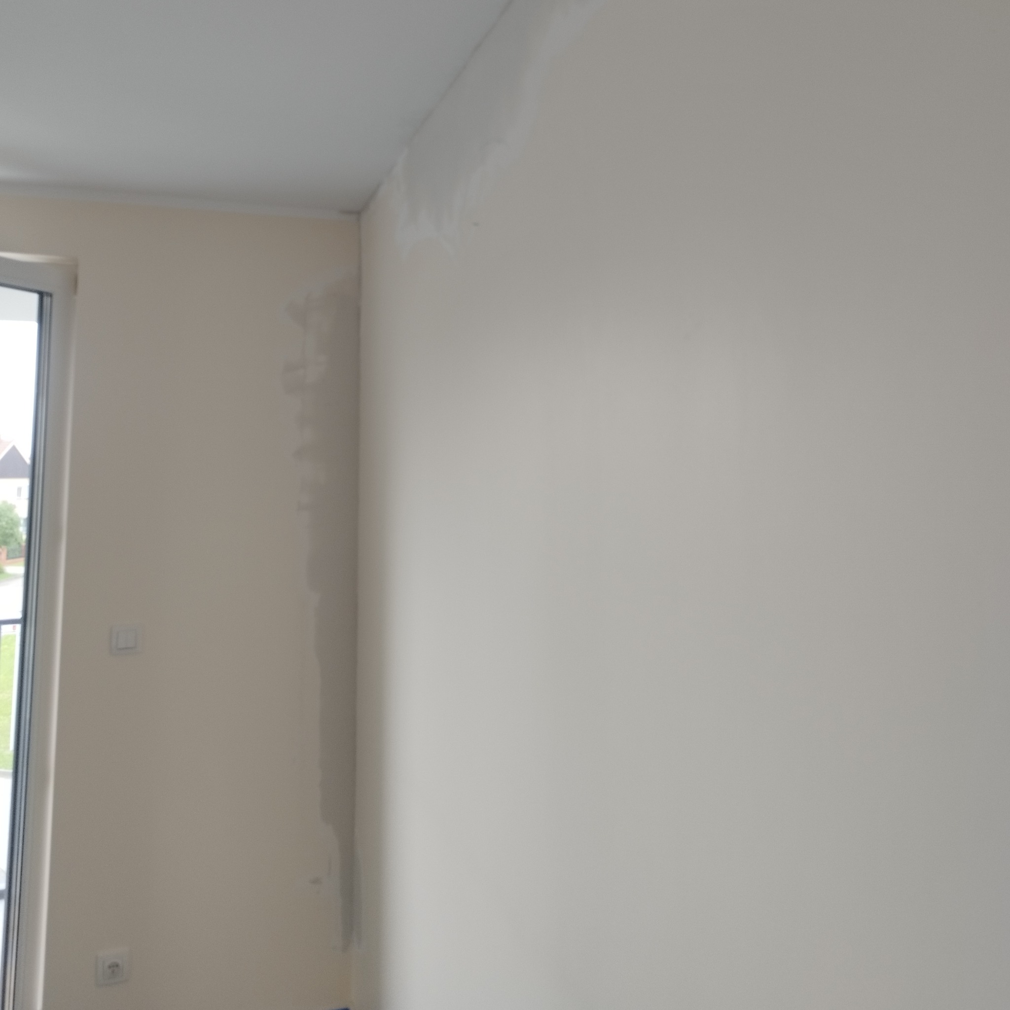 Ściana wewnętrzna w trakcie prac przygotowawczych do malowania, widoczne ślady po szpachlowaniu i nierówności, okno z widokiem na zieleń na zewnątrz.