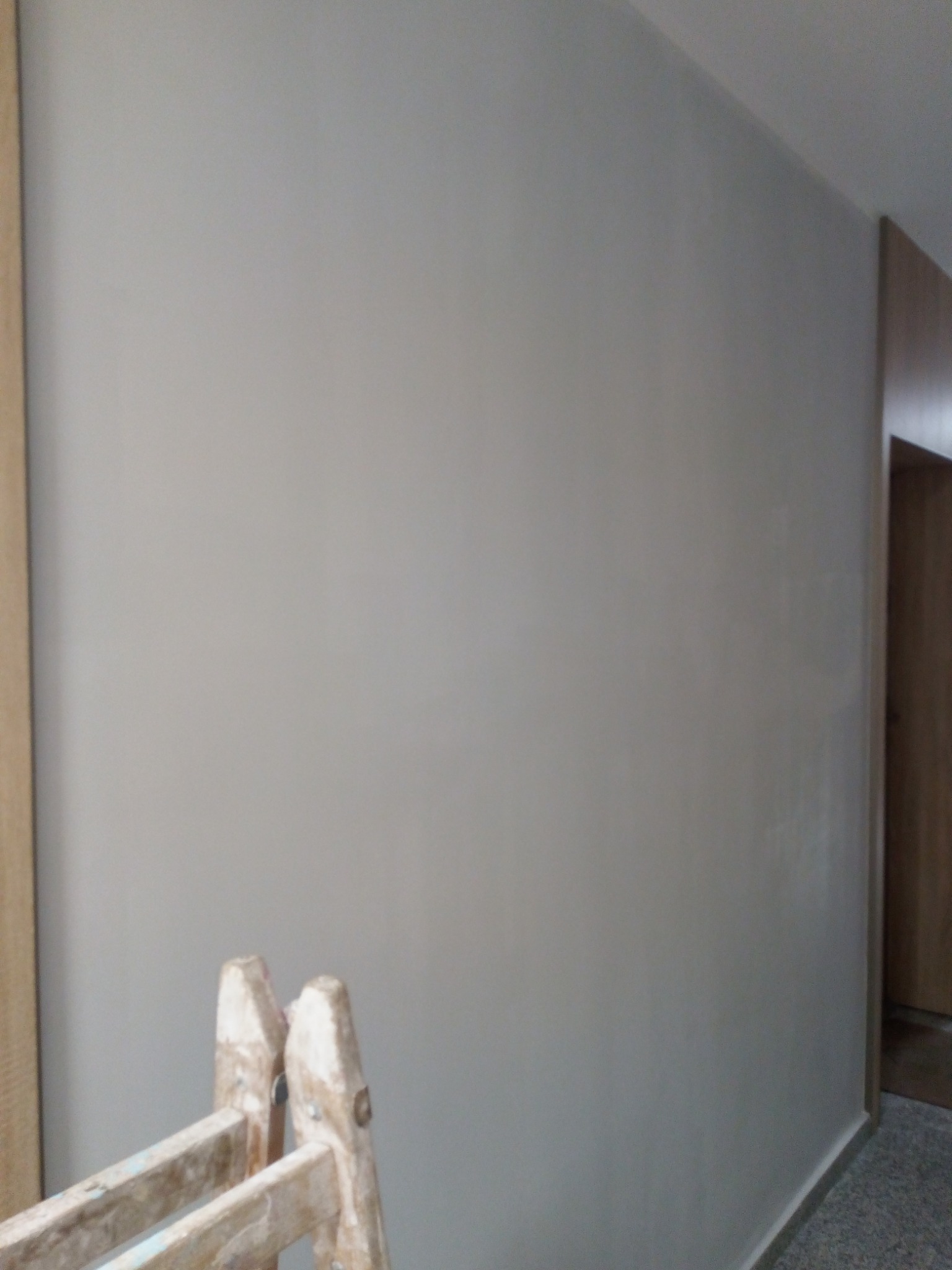 Świeżo pomalowana ściana w odcieniach szarości, z widoczną starą, drewnianą drabiną malarską w dolnej części kadru, sugerująca etap prac wykończeniowych.