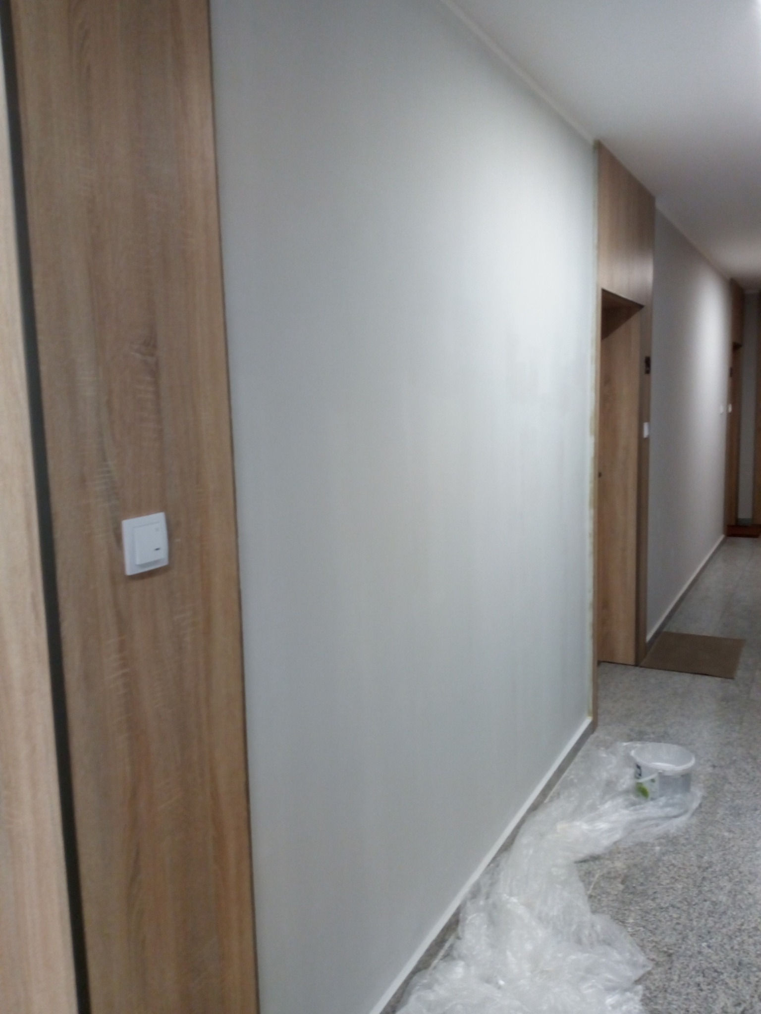 Świeżo pomalowana ściana w korytarzu z widoczną osłoną z folii malarskiej na podłodze i wiadrem po farbie, obok drewnianej futryny drzwi z włącznikiem światła.