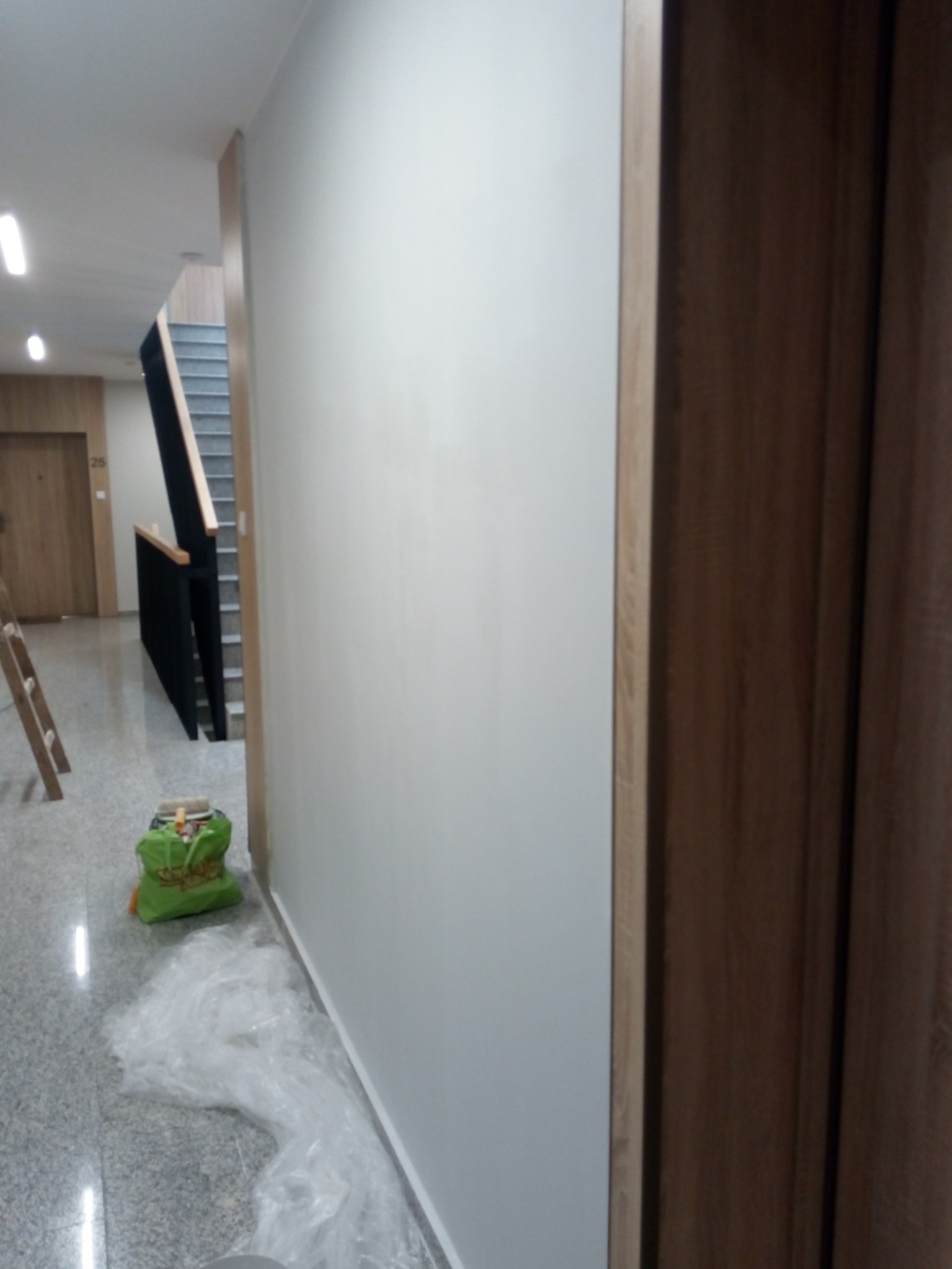 Świeżo pomalowana, gładka, szara ściana w korytarzu, widoczny fragment drewnianej ościeżnicy drzwi, zabezpieczenia podłogi folią malarską oraz torba z narzędziami malarskimi w tle.