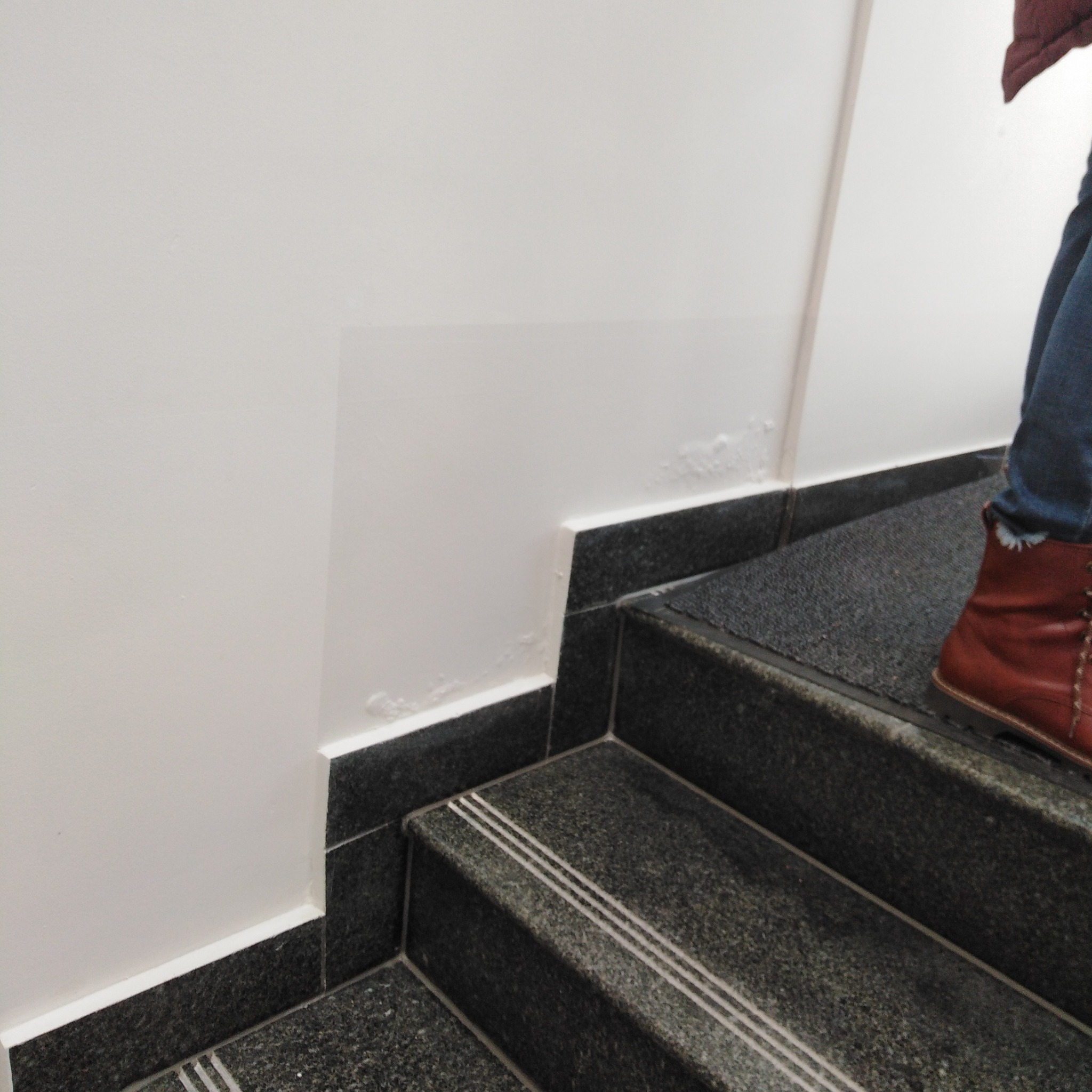 Fragment schodów z granitu z trzema białymi paskami antypoślizgowymi, dolna część ściany po świeżym malowaniu, widoczne niedoskonałości przy listwie przypodłogowej, fragment nogi w jeansach...