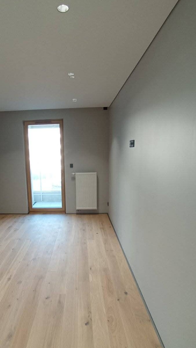 Wnętrze po odświeżeniu: gładkie, szare ściany, jasna drewniana podłoga, biały grzejnik pod oknem, widoczne gniazdka elektryczne.