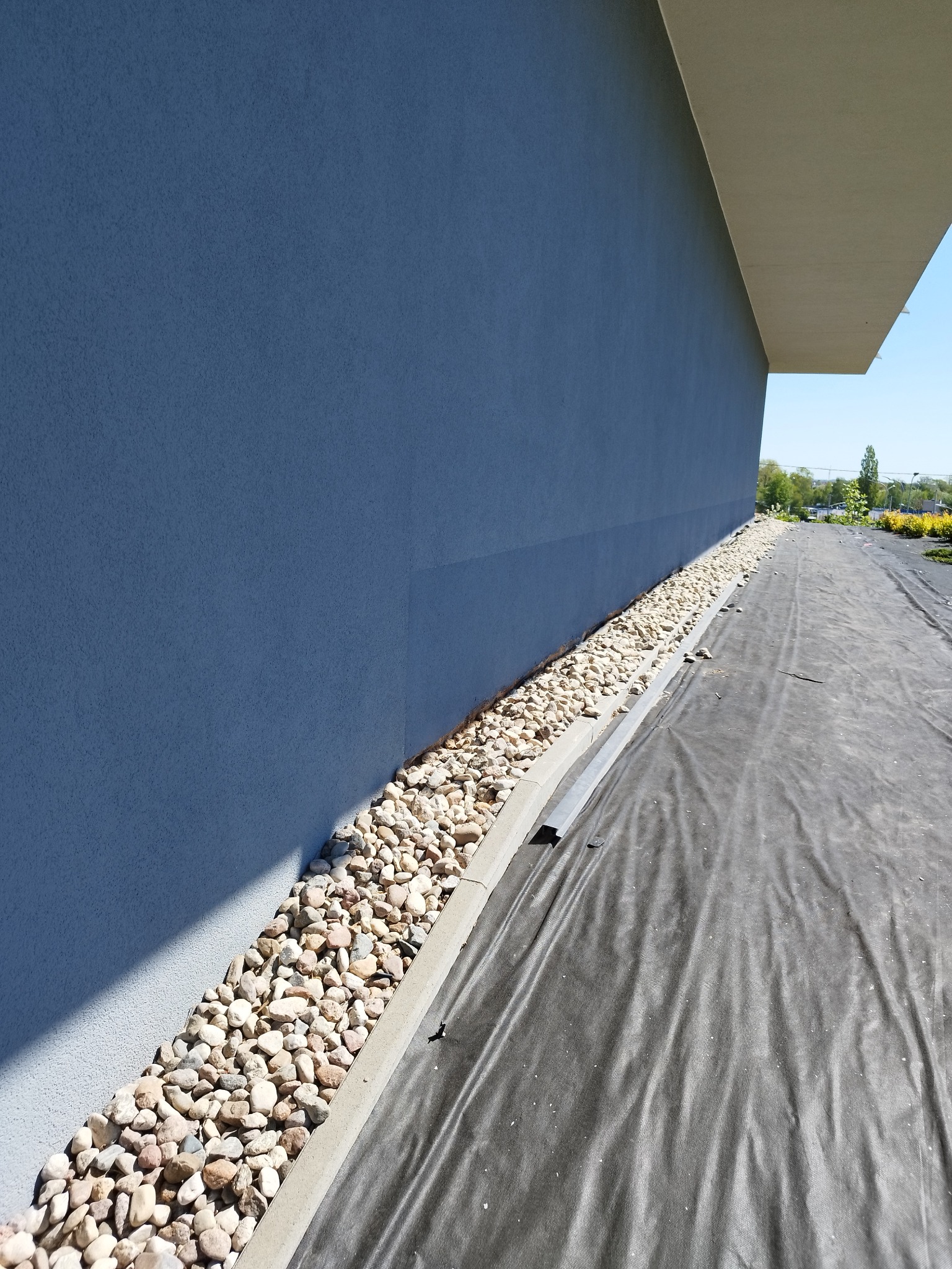 Niebieska elewacja budynku z wykończeniem z jasnych kamieni oddzielonych betonowym obrzeżem, widoczna czarna folia ochronna na ziemi.