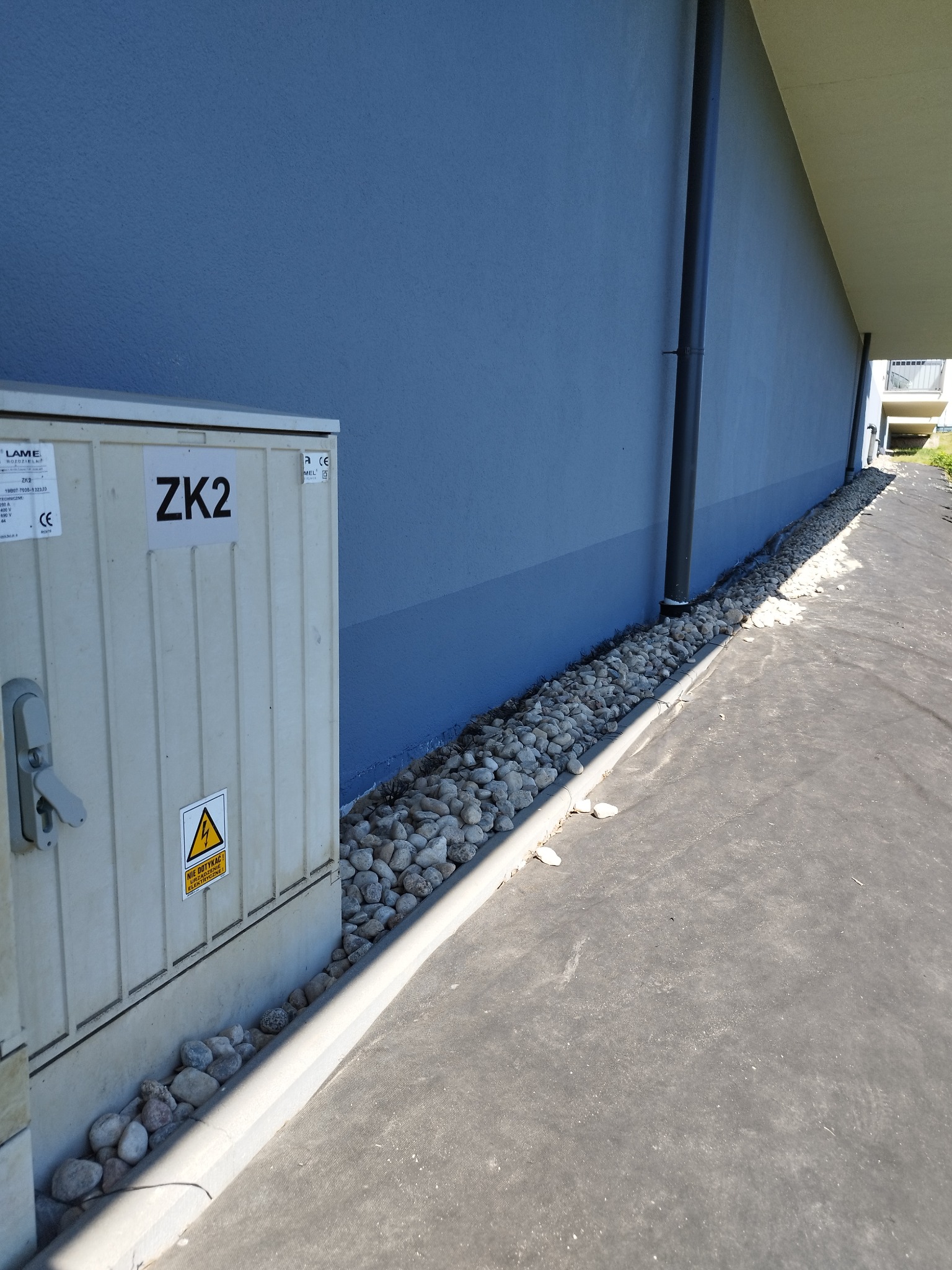 Niebieska ściana budynku z rurą spustową i skrzynką elektryczną ZK2, obłożona kamieniami, widok z boku.