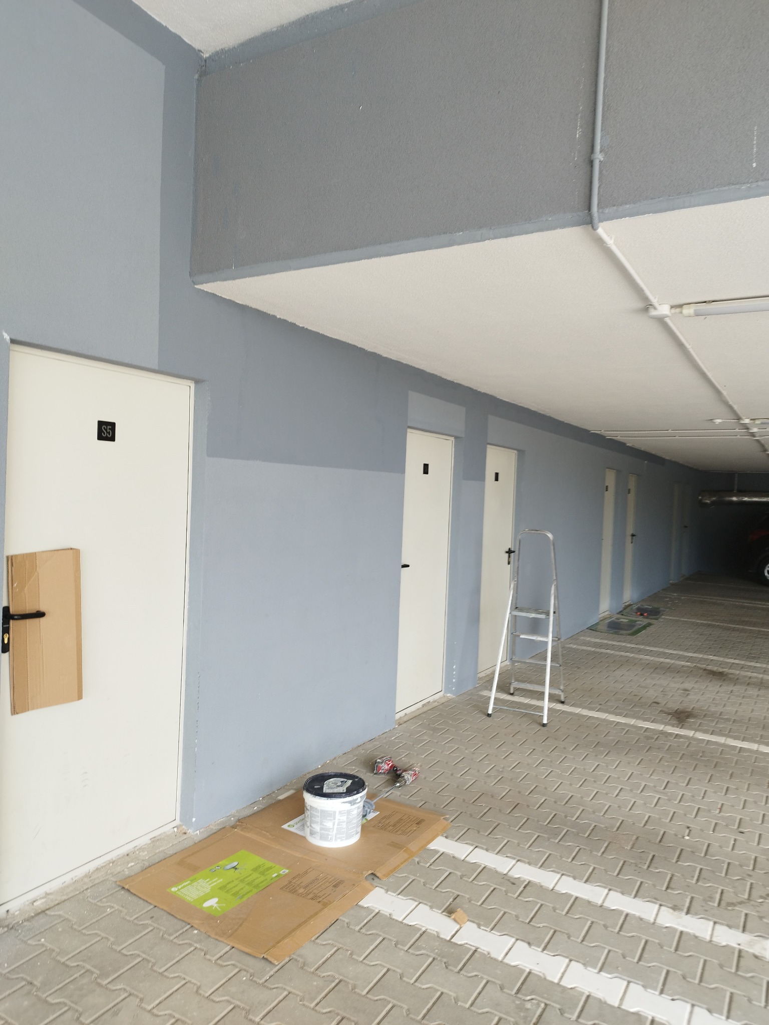 Korytarz garażowy z pomalowanymi ścianami na szaro-niebieski kolor, białe drzwi z numerami, drabina aluminiowa, puszka farby na kartonie i fragment zabezpieczonej kartonem klamki.