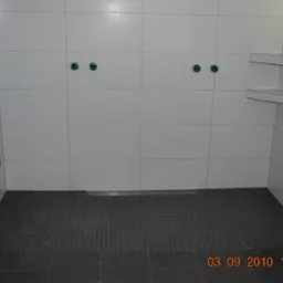 Wnętrze łazienki w trakcie remontu: białe kwadratowe płytki na ścianach z widocznymi otworami na armaturę, szare mozaikowe płytki podłogowe i odpływ liniowy.