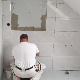 STASZEK GLAZURNIK - Glazurnik w trakcie pracy w Piaskach: Układanie białych, błyszczących płytek w łazience na poddaszu, widoczny fragment ściany z zaznaczonym miejscem na lustro i wyprowadzonymi kablami, oraz wiadro...