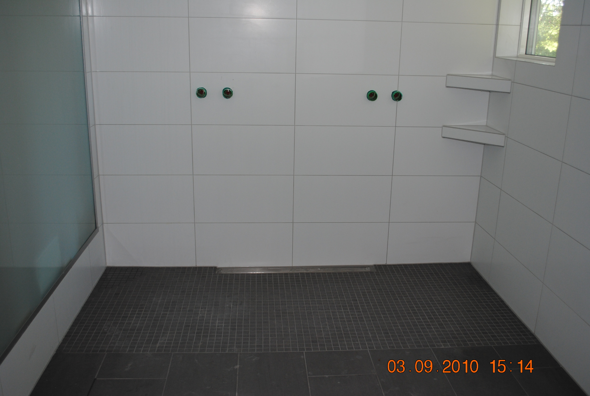 Wnętrze łazienki w trakcie remontu: białe kwadratowe płytki na ścianach z widocznymi otworami na armaturę, szare mozaikowe płytki podłogowe i odpływ liniowy.