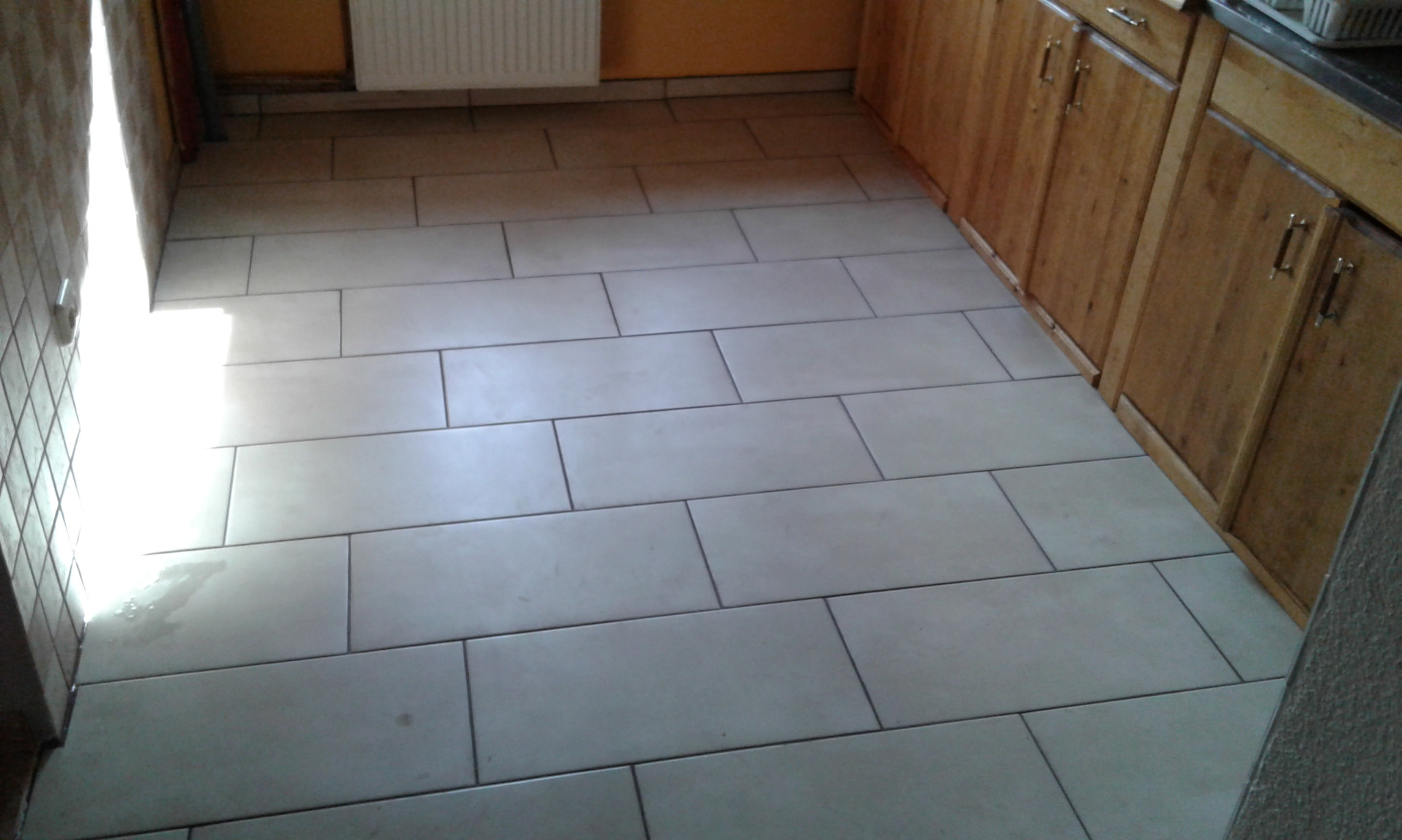 Ułożona podłoga z jasnych, prostokątnych płytek w kuchni, widoczne drewniane szafki kuchenne i fragment ściany z grzejnikiem.