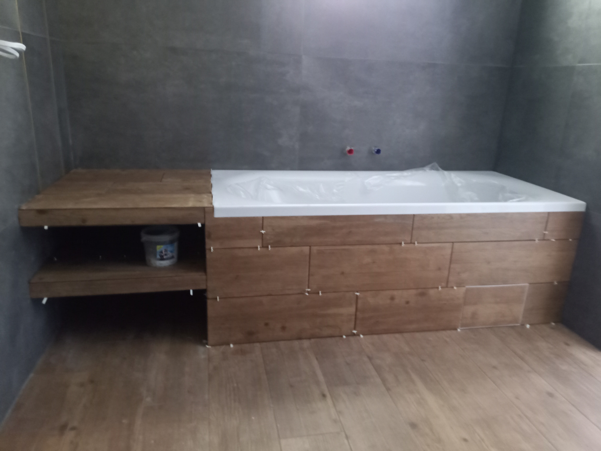 Obudowana wanna z drewnopodobnymi płytkami w trakcie prac wykończeniowych, widoczny pojemnik z zaprawą na półce obok.