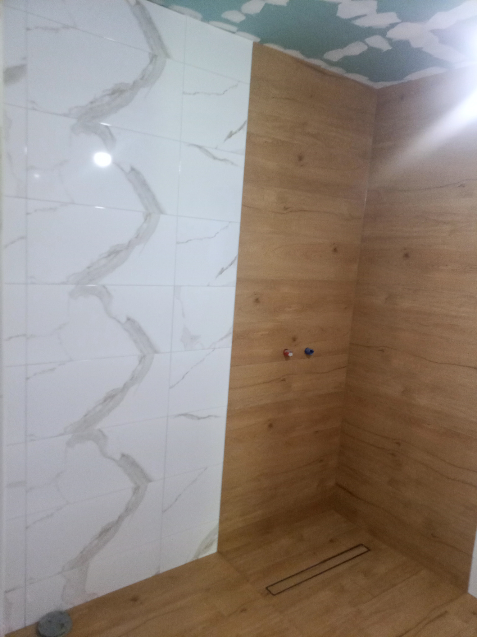 Fragment wnętrza łazienki w trakcie prac wykończeniowych, z widocznymi płytkami imitującymi marmur na jednej ścianie i drewno na drugiej, z zamontowaną podłogą i odpływem liniowym, sufit w stanie...