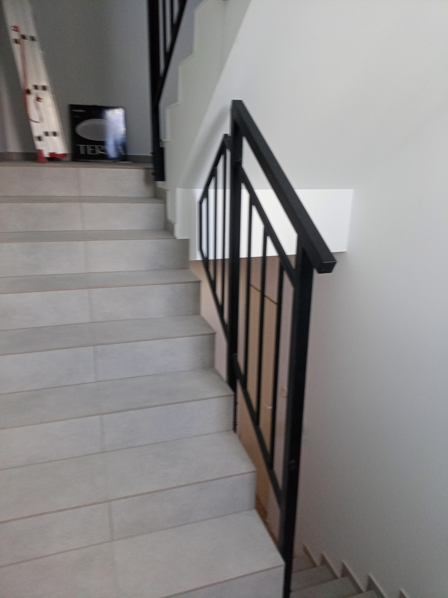 Schody wyłożone płytkami ceramicznymi w odcieniach szarości, z czarną metalową balustradą, widok z dołu. Na górze schodów oparta o ścianę biała drabina aluminiowa i czarne pudełko z napisem.