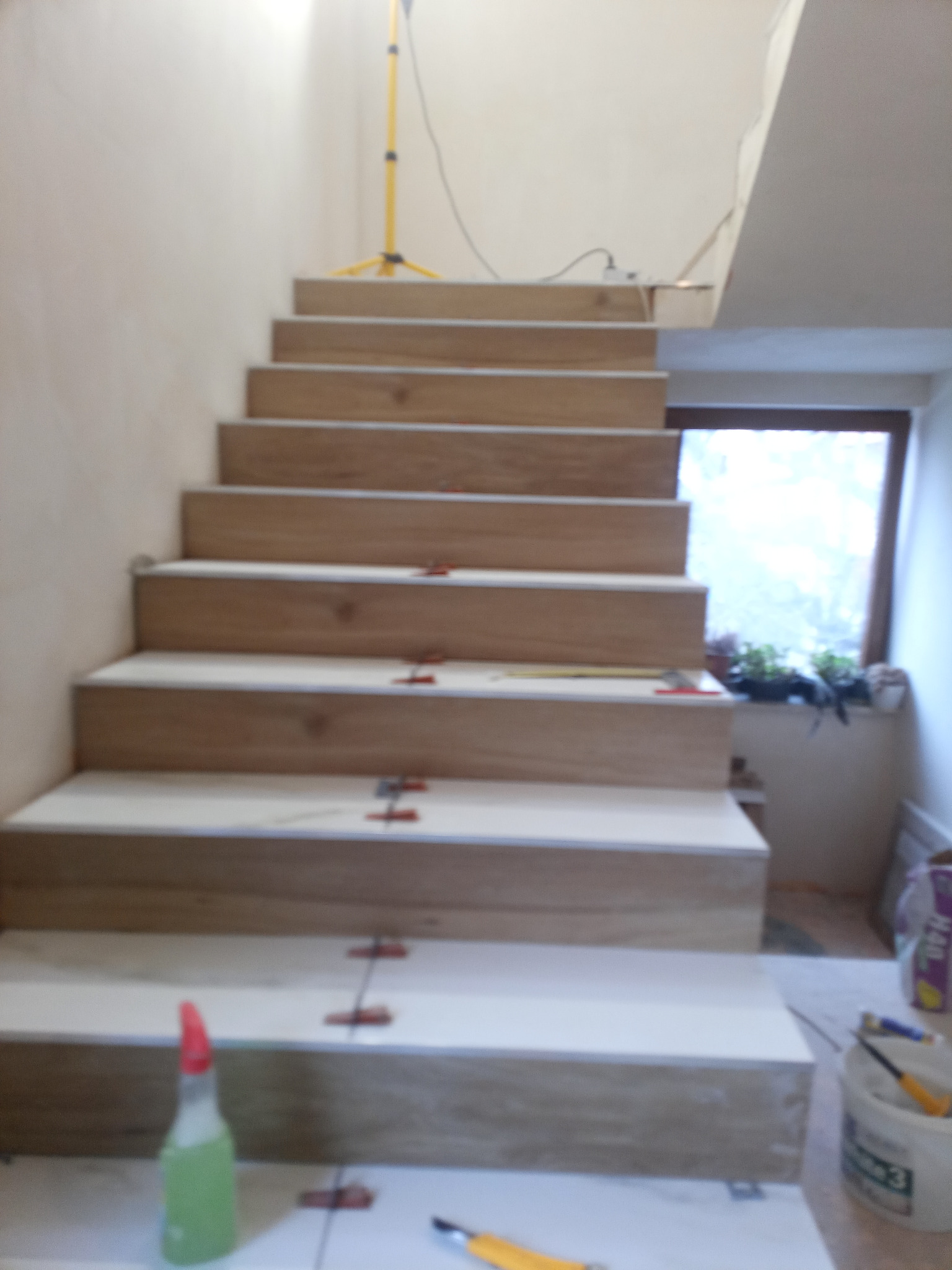 Schody w trakcie układania płytek imitujących drewno, widoczne krzyżyki dystansowe, butelka ze spryskiwaczem i lampa budowlana w tle.