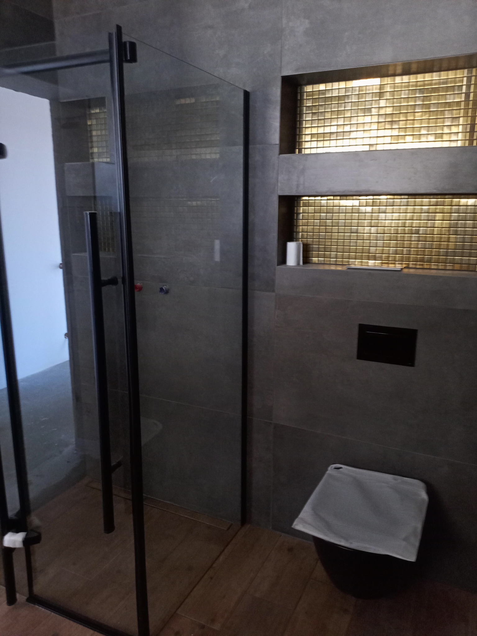 Nowoczesna łazienka z szarą, teksturowaną ścianą, złotą mozaiką w niszach, kabiną prysznicową z czarnymi ramami i drewnopodobną podłogą; na ścianie widoczny czarny panel sterujący, a na półce rolka...