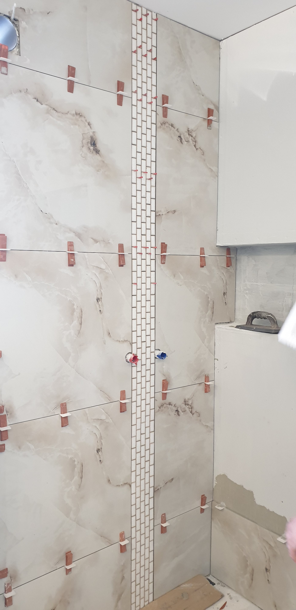 Układanie płytek imitujących marmur z dekoracyjnym pasem małych, białych kafelków w łazience, z użyciem klipsów poziomujących.