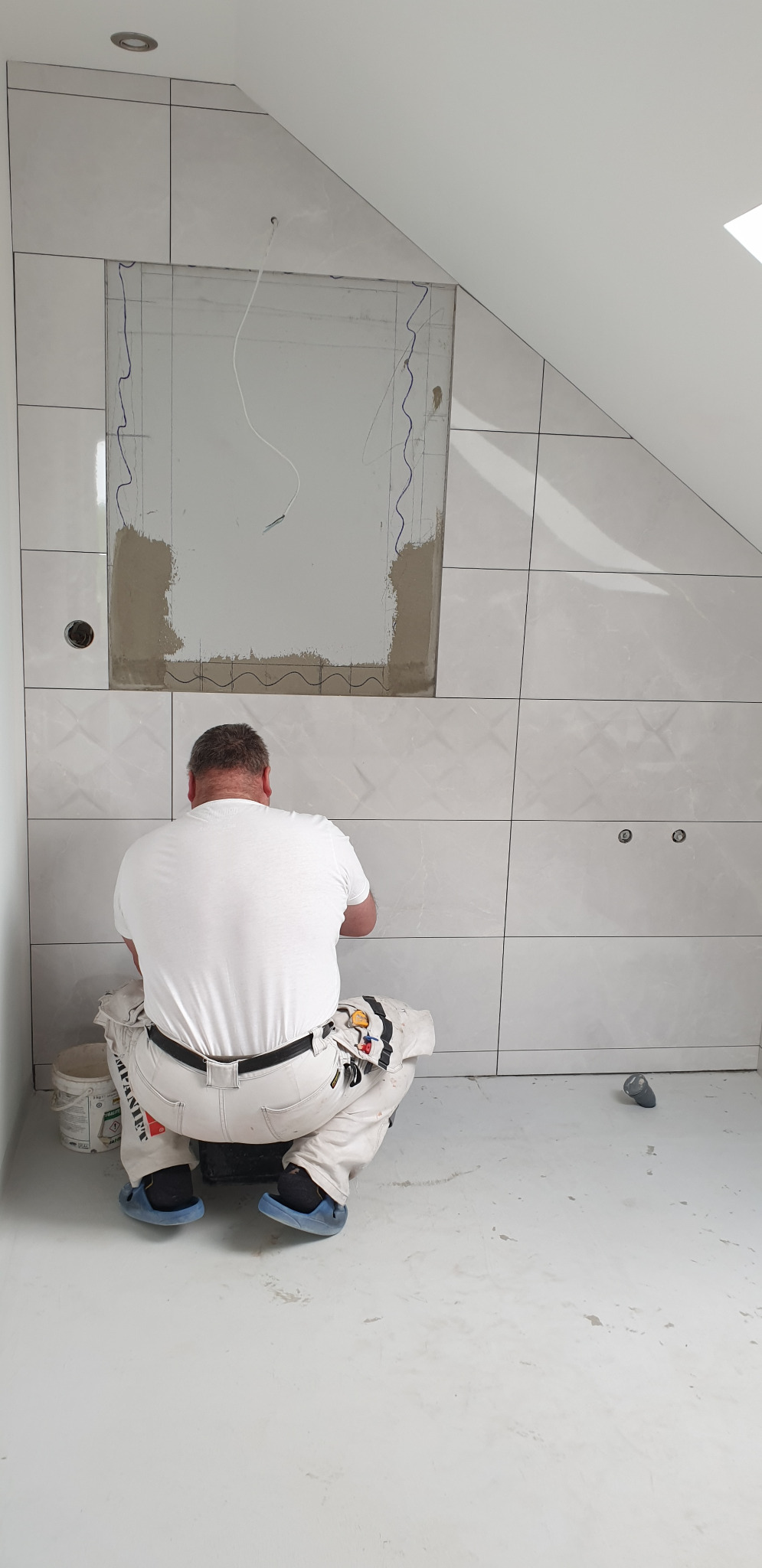 Glazurnik w trakcie pracy w Piaskach: Układanie białych, błyszczących płytek w łazience na poddaszu, widoczny fragment ściany z zaznaczonym miejscem na lustro i wyprowadzonymi kablami, oraz wiadro...