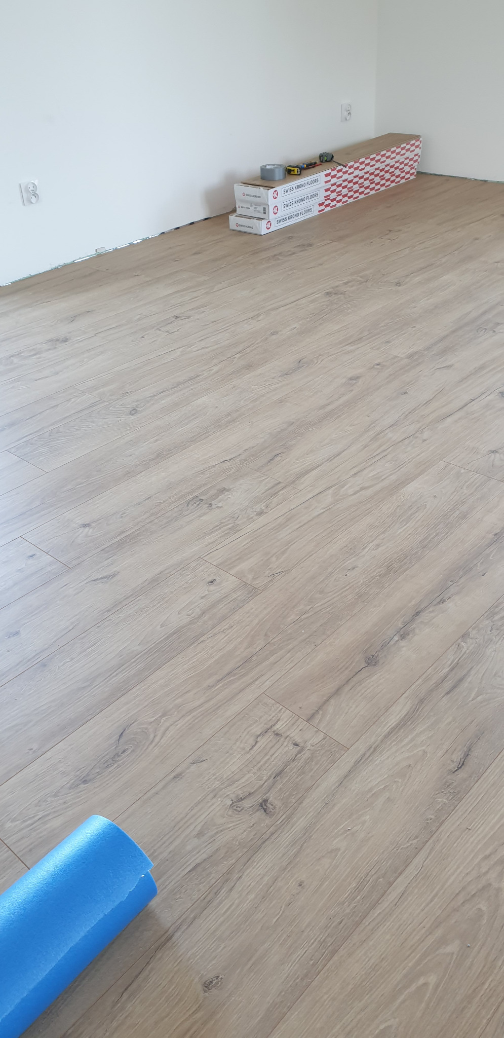 Jasne panele podłogowe ułożone w pokoju, w tle kartony paneli Swiss Krono Floors, taśma klejąca i narzędzia, niebieska pianka podkładowa w rogu.