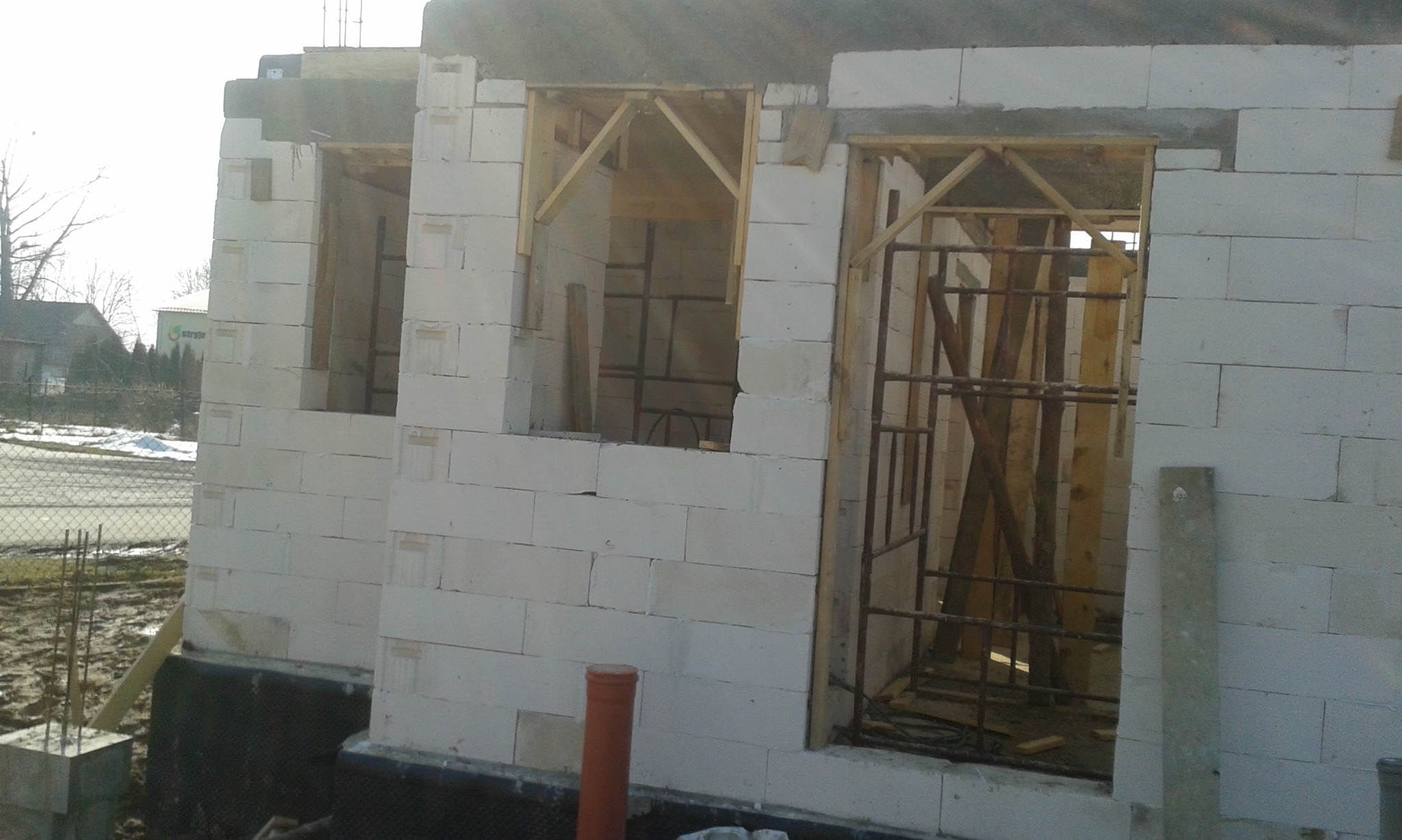 Surowy stan budynku w trakcie wznoszenia ścian z betonu komórkowego, widoczne otwory okienne i drzwiowe z drewnianym szalunkiem i stalowymi wzmocnieniami.