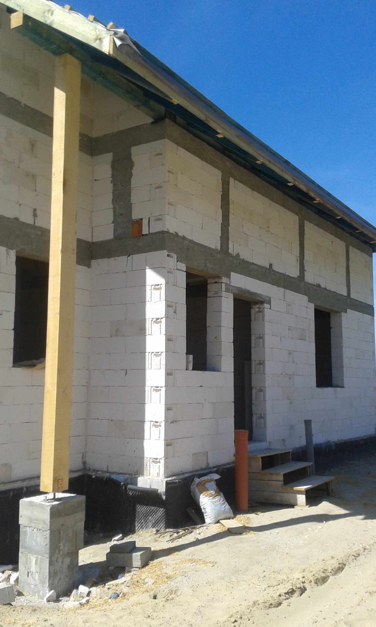 Surowy stan budynku z białych bloczków, widoczne otwory okienne i drzwiowe, pionowa drewniana podpora, fundament zabezpieczony hydroizolacją, worek cementu przy schodach wejściowych.