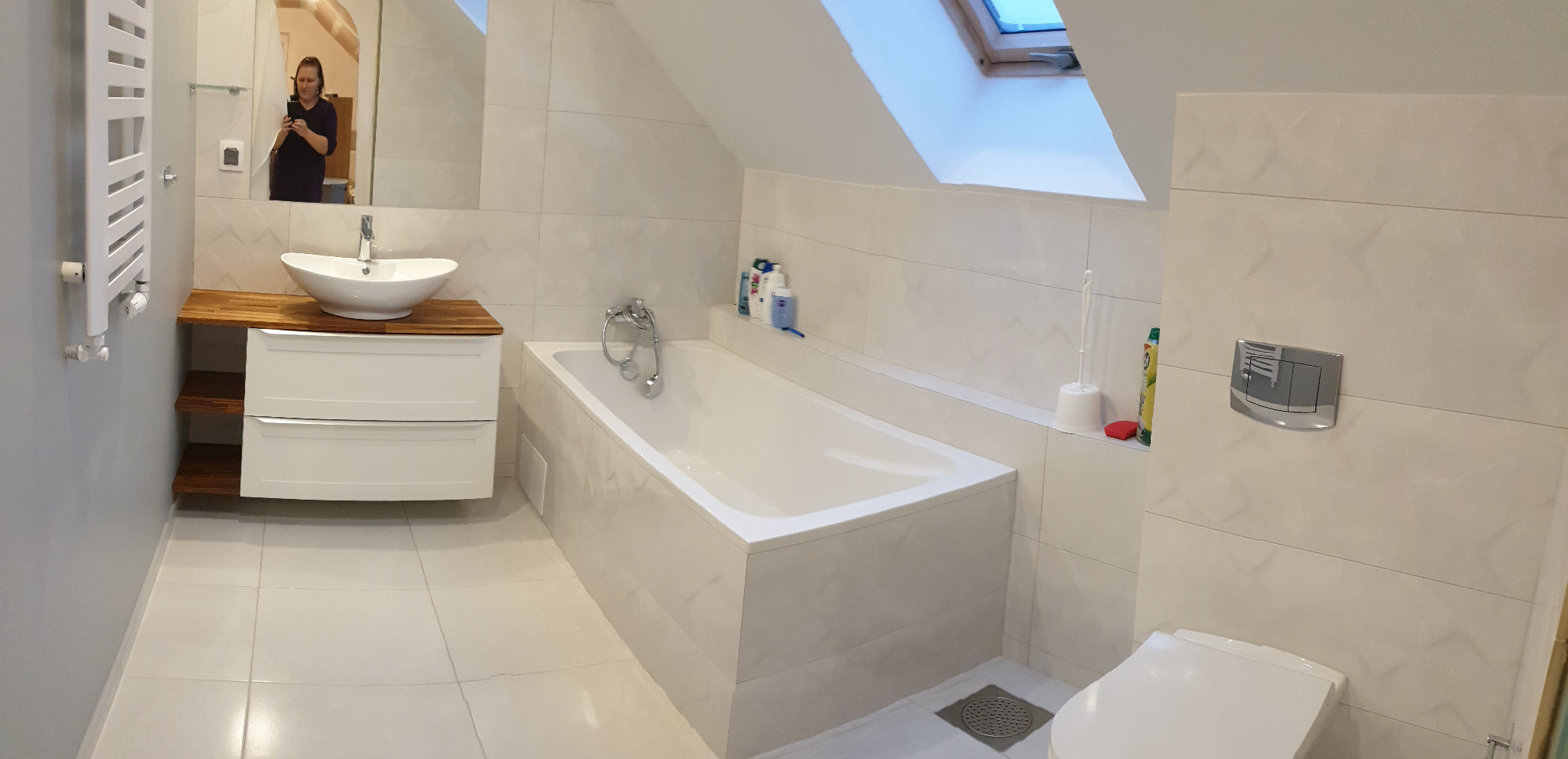 Jasna łazienka na poddaszu z białą wanną, umywalką na drewnianym blacie i toaletą w zabudowie, sfotografowana z perspektywy osoby stojącej w wejściu.