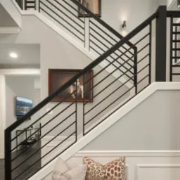 Nowoczesna balustrada schodowa z czarnych, poziomych prętów, uzupełniona dekoracyjnymi poduszkami na drewnianej ławce u podnóża schodów, z obrazami w ramkach na ścianie.