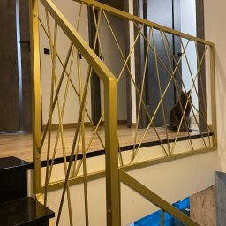 Loftski Poznań bramy przęsła balustrady - Złota balustrada wewnętrzna z geometrycznym wzorem, przez którą widać siedzącego kota rasy Maine Coon, z czarnymi schodami i fragmentem turkusowej sofy w tle.
