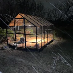 Szklarnia z czarną metalową konstrukcją, oświetlona od wewnątrz, widoczna podczas śnieżnej nocy, z narzędziami i materiałami budowlanymi na zewnątrz i wewnątrz.