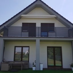 Loftski Poznań bramy przęsła balustrady - Nowoczesny dom z szarą, poziomą balustradą na balkonie, widok z ogrodu na elewację z dwoma oknami i tarasem z meblami ogrodowymi.