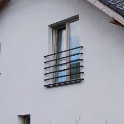 Loftski Poznań bramy przęsła balustrady - Okienko z czarną, poziomą balustradą z prostokątnych profili na białej elewacji domu z szarym dachem.