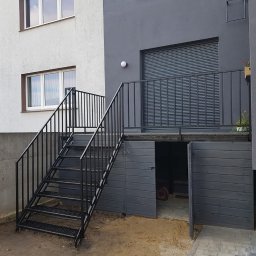 Loftski Poznań bramy przęsła balustrady - Zewnętrzne schody metalowe z czarną balustradą prowadzące do podwyższonego tarasu z szarą roletą i wejściem do garażu, widok na dwa budynki o różnej kolorystyce elewacji.