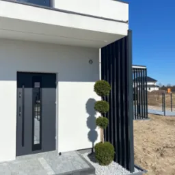 Nowoczesny dom jednorodzinny z antracytowymi elementami: drzwi wejściowe, pionowe panele dekoracyjne oraz kuliste krzewy na tle białej elewacji i kostki brukowej.