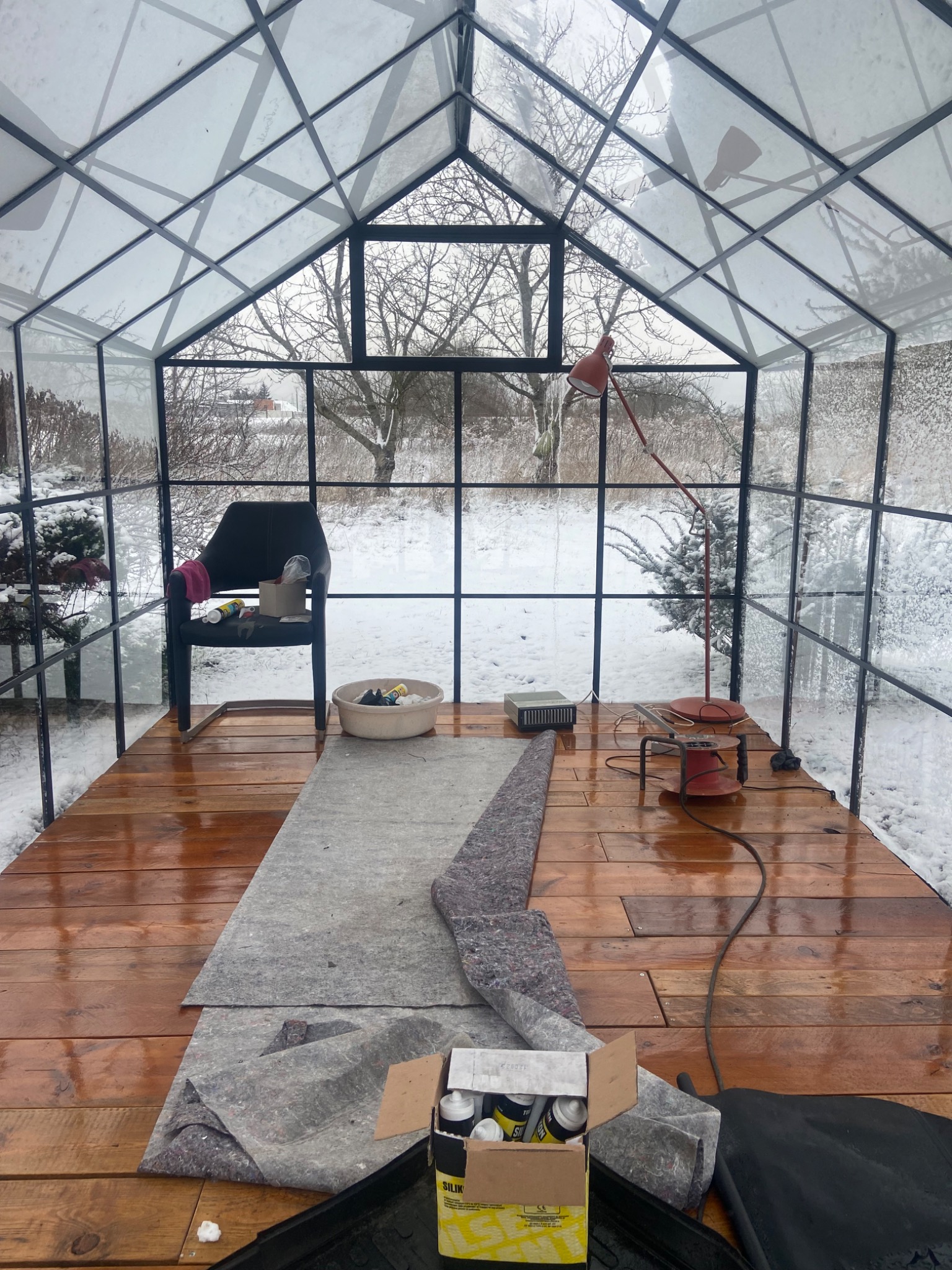 Wnętrze przeszklonej konstrukcji z drewnianą podłogą w trakcie prac wykończeniowych, widoczne narzędzia, materiały izolacyjne i lampa na tle zimowego krajobrazu za oknem.