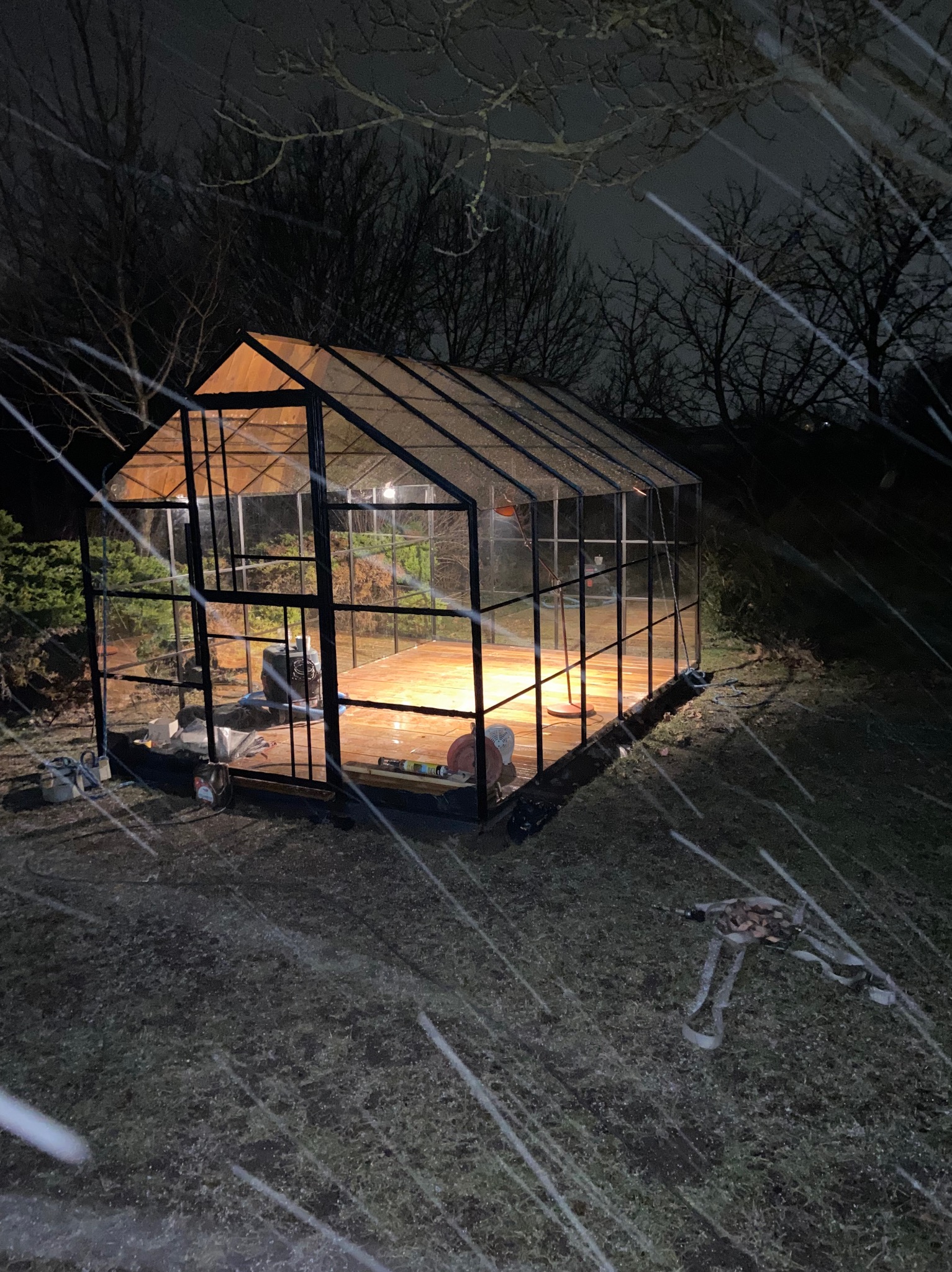 Szklarnia z czarną metalową konstrukcją, oświetlona od wewnątrz, widoczna podczas śnieżnej nocy, z narzędziami i materiałami budowlanymi na zewnątrz i wewnątrz.