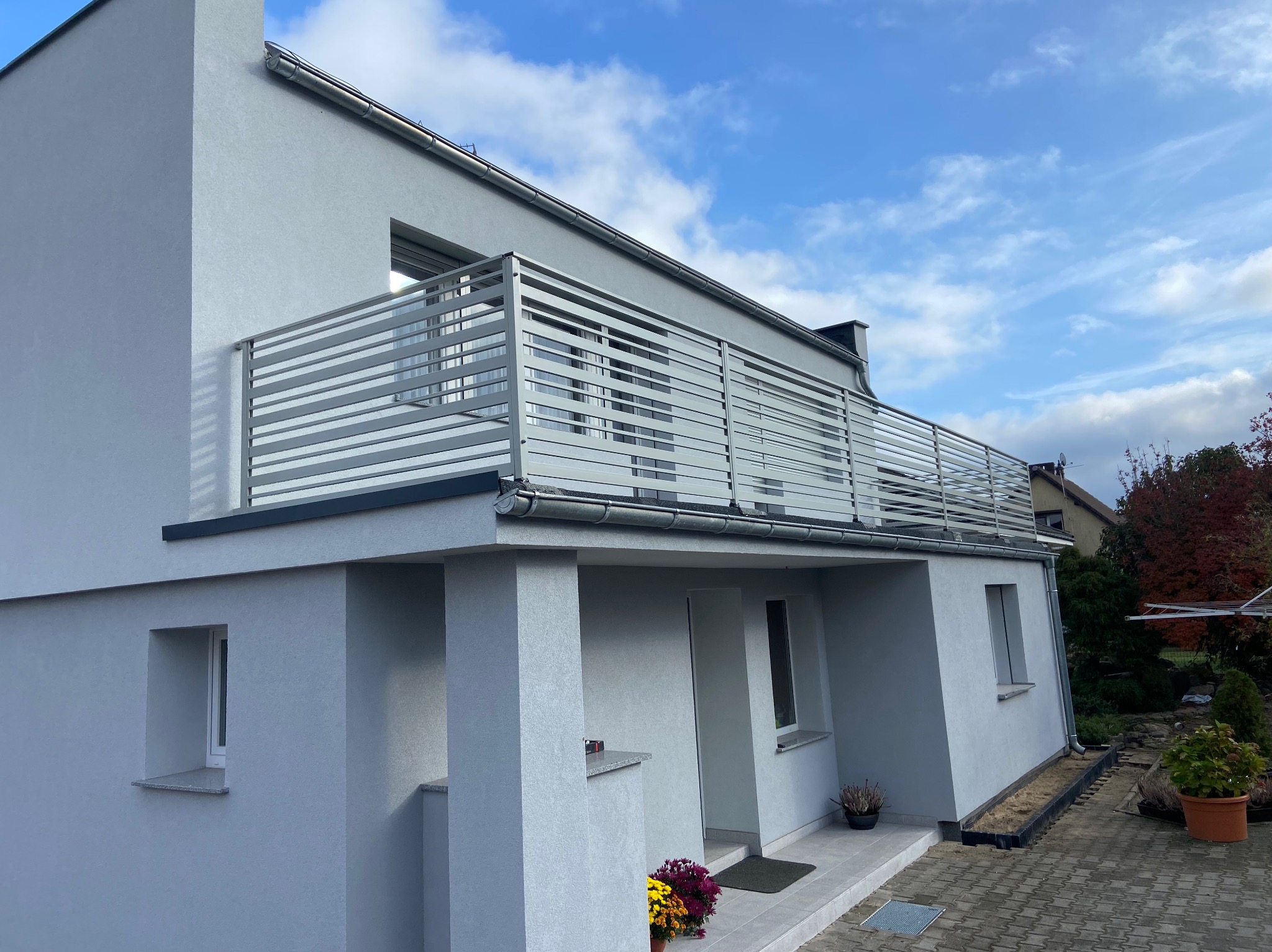 Nowoczesny dom z szarą elewacją i balkonem z balustradą nierdzewną o poziomych szczeblach, widok z zewnątrz, na pierwszym planie wejście do domu z donicami kwiatów.