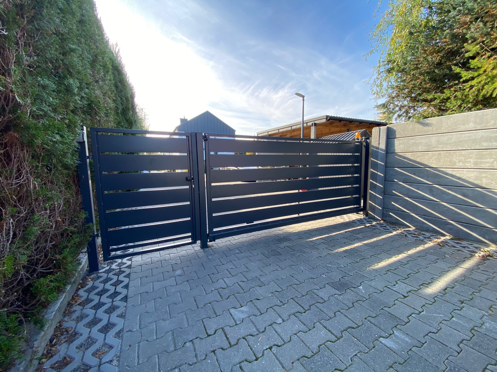 Nowoczesna, antracytowa brama wjazdowa z poziomymi panelami, częściowo otwarta, z betonowym ogrodzeniem po prawej stronie i żywopłotem po lewej, na tle błękitnego nieba.