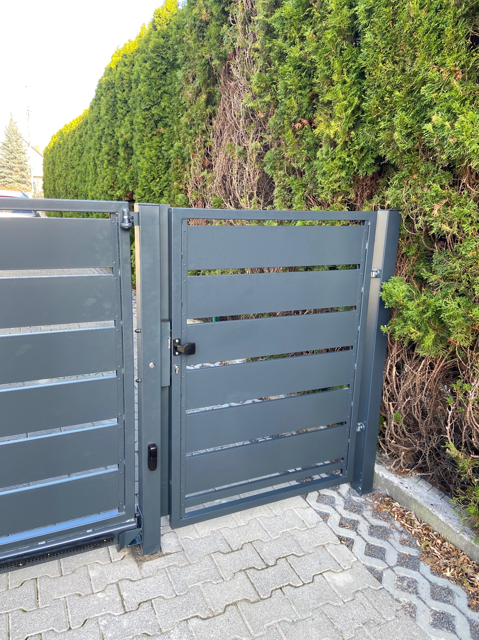 Nowoczesna, szara brama wjazdowa z poziomymi panelami, automat do bramy widoczny u dołu, na tle żywopłotu i kostki brukowej.