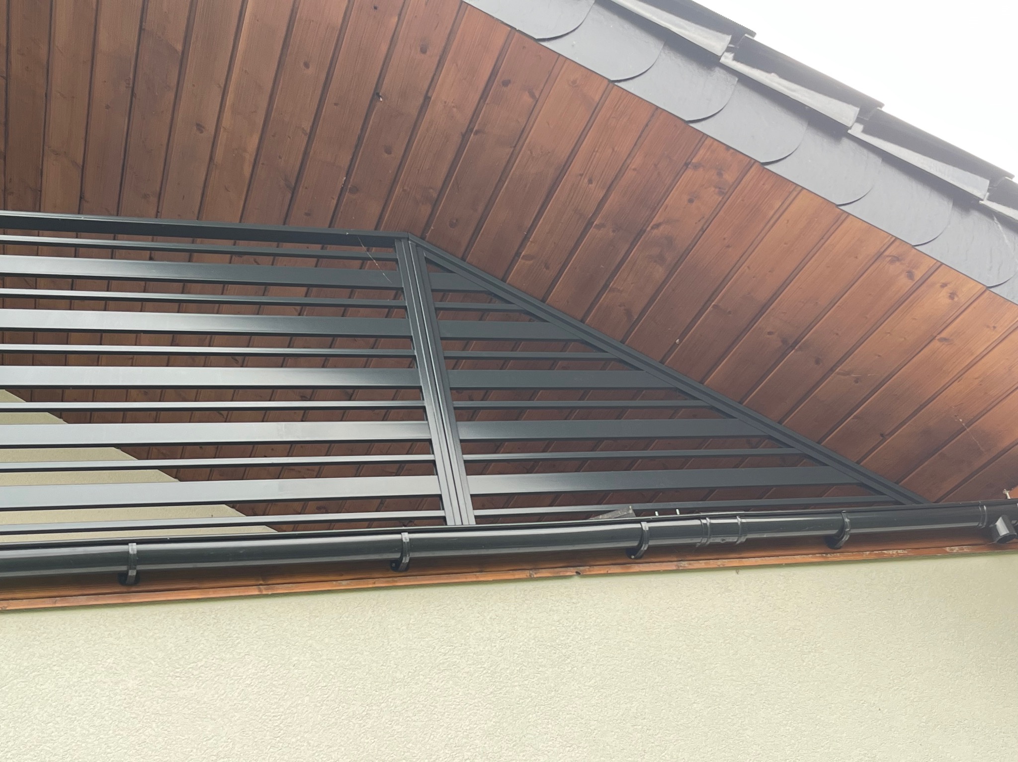 Metalowa pergola z poziomymi listwami zamontowana pod okapem dachu z drewnianą podbitką i rynną, widok z dołu.