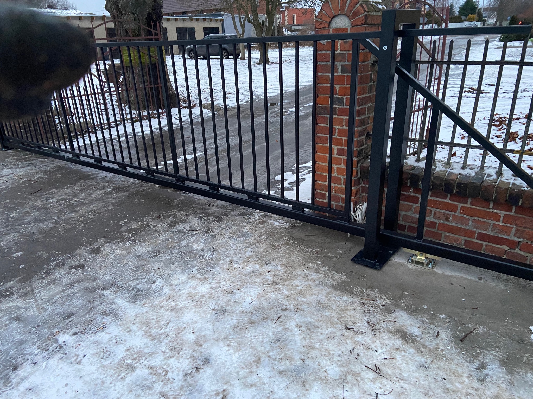Czarna, przesuwna brama wjazdowa z metalowymi prętami, zamontowana na ceglanym słupku, widoczny mechanizm jezdny na betonowej nawierzchni pokrytej śniegiem, w tle budynki i drzewa.