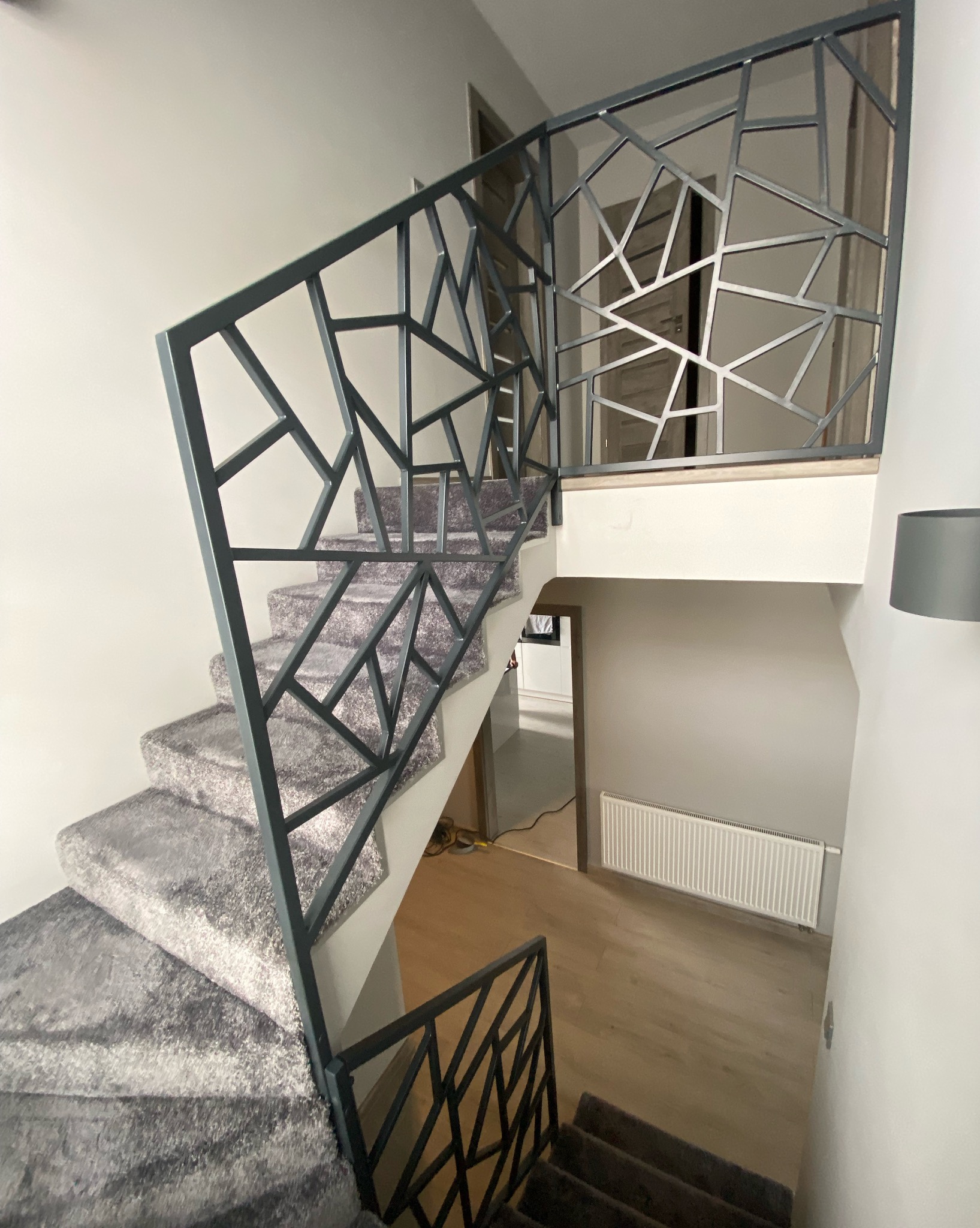 Nowoczesna balustrada schodowa o geometrycznym wzorze, wykonana z metalu w odcieniach szarości, na tle schodów pokrytych szarym dywanem.