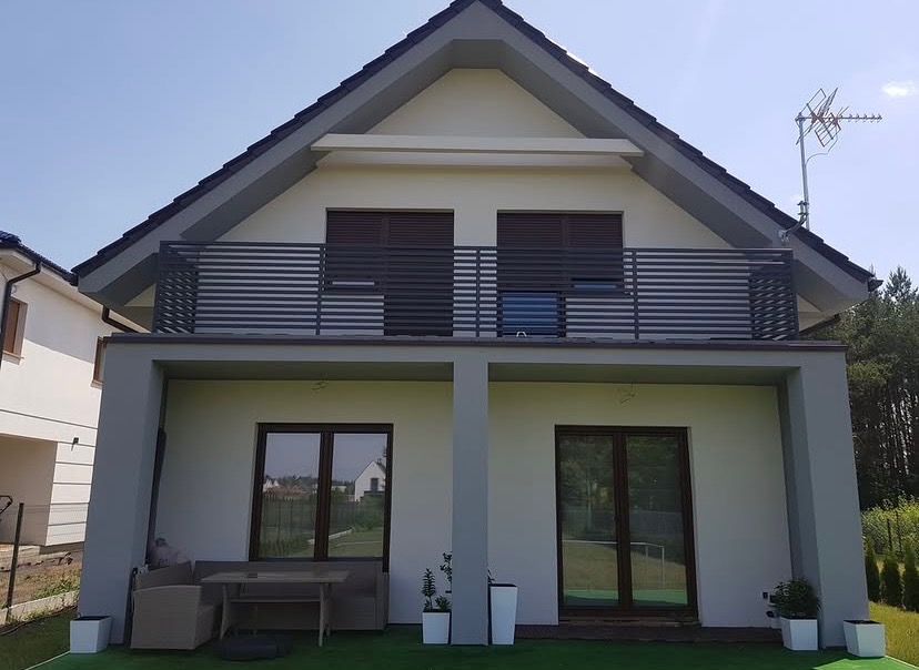 Nowoczesny dom z szarą, poziomą balustradą na balkonie, widok z ogrodu na elewację z dwoma oknami i tarasem z meblami ogrodowymi.