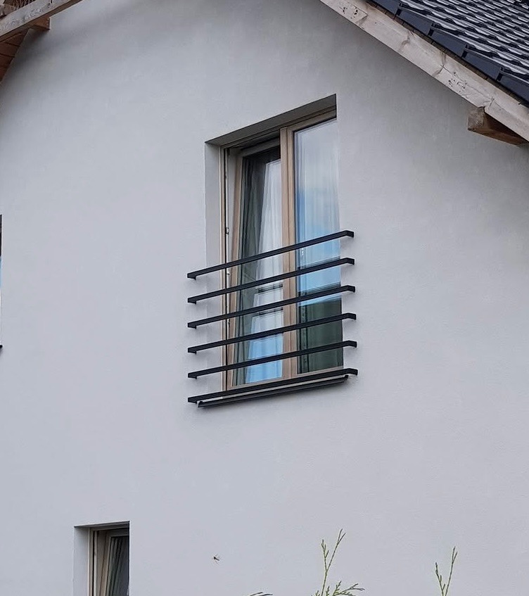 Okienko z czarną, poziomą balustradą z prostokątnych profili na białej elewacji domu z szarym dachem.