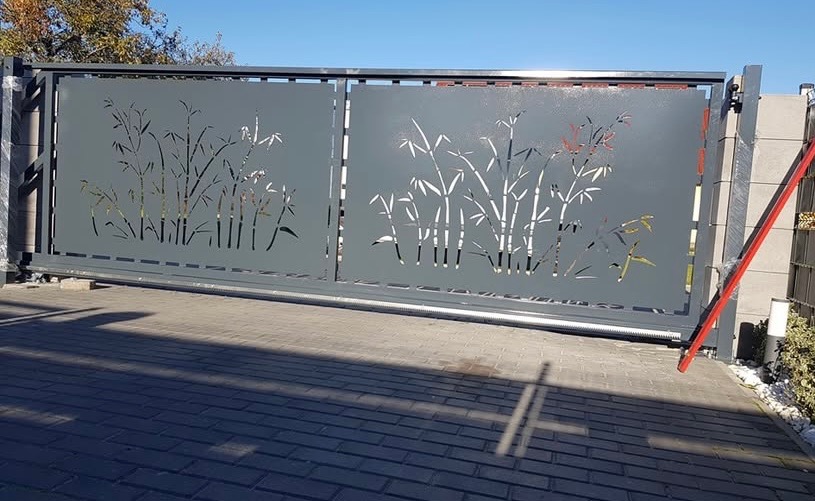 Przesuwna brama wjazdowa z szarego metalu, zdobiona wycinanym motywem bambusa, zamontowana na posesji z brukowanym podjazdem.