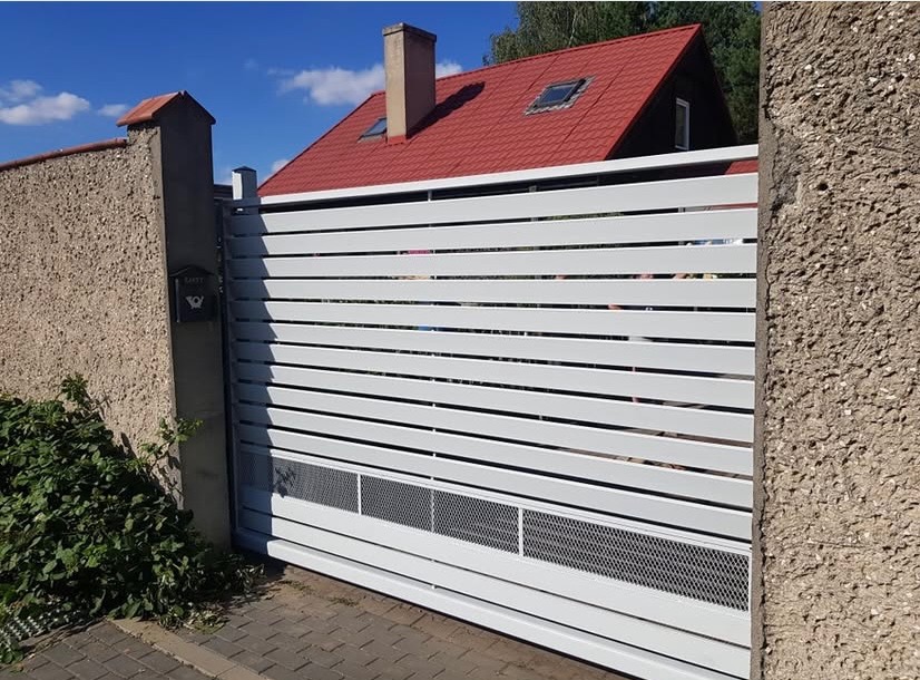 Nowoczesna, przesuwna brama wjazdowa z poziomych, białych profili, z ażurowym panelem wentylacyjnym u dołu, zamontowana między betonowymi słupkami z czarną skrzynką na listy