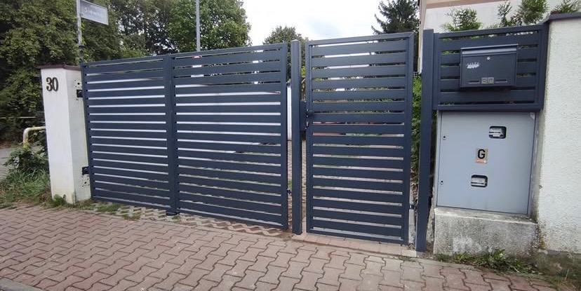 Nowoczesna brama wjazdowa i furtka z poziomych, grafitowych profili, skrzynka na listy zintegrowana z jednym ze słupków, kostka brukowa na podjeździe.