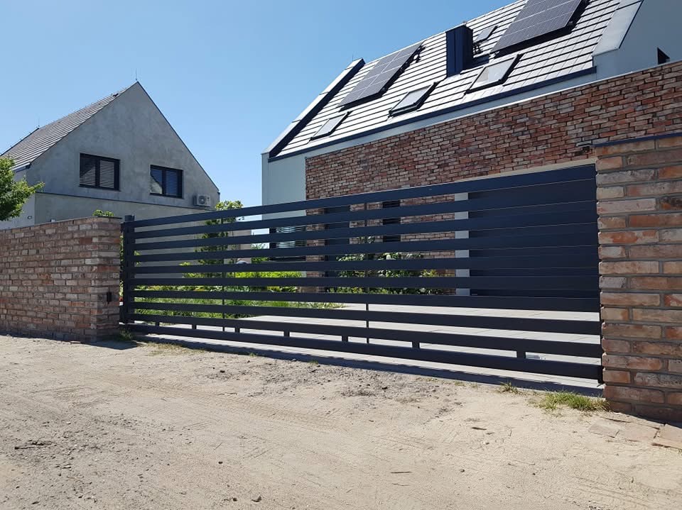 Nowoczesna, pozioma brama wjazdowa z metalowych profili, w kolorze grafitowym, osadzona między murkami z cegły klinkierowej, na tle dwóch budynków mieszkalnych, jeden z panelami solarnymi na dachu.